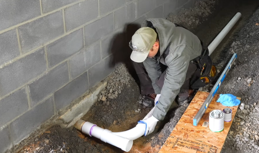 Installing a new PVC drain line in an excavated trench beside a foundation wall